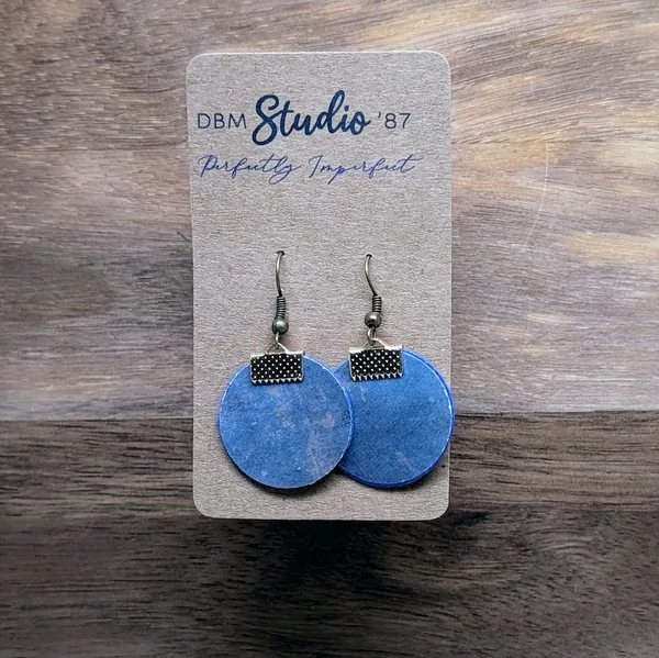 quarter size round earrings made from layers of paper modpodged together, blue with swirls of gold/copper front and beige with white cursive back. The color of the sides made by the paper inside is blue. Brass-tone findings.